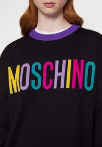 Femme aux cheveux rouges portant un pull noir avec un col violet et le mot « MOSCHINO » en lettres colorées sur la poitrine.