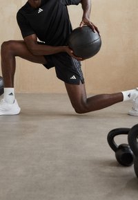 Svart atletisk skjorta och shorts med vita Adidas-stripes, håller en svart medicinboll. Vita sneakers och kettlebells i förgrunden.