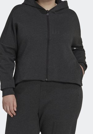 Osoba nosząca ciemnoszarą bluzę rozpinaną na zamek z kapturem i pasujące spodnie dresowe z logo Adidas na lewym rękawie i nogawkach.