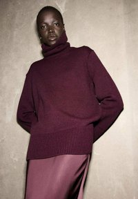 Maglione a collo alto bordeaux con texture a costine e orlo, indossato sopra una gonna lunga bordeaux liscia, su uno sfondo neutro.