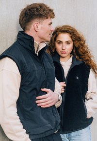 Fekete fleece mellény magas gallérral, cipzárral és logóval; bézs réteges pulóver alatta; laza farmernadrág.