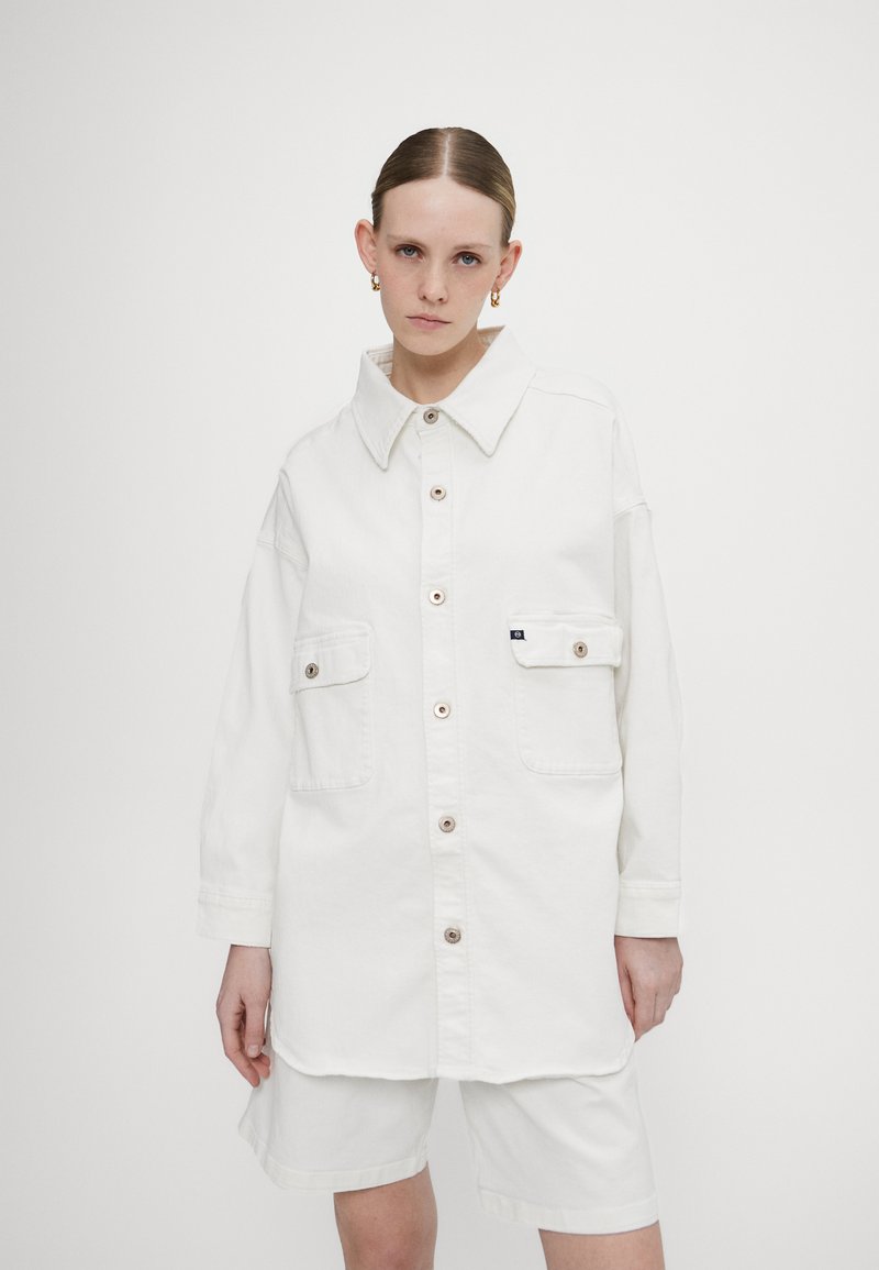 Giacca camicia in denim bianco con colletto, chiusura frontale con bottoni, due tasche sul petto e maniche lunghe. Texture liscia, vestibilità rilassata e branding discreto.