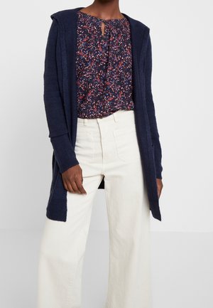 Femme portant un cardigan bleu marine ouvert, un chemisier à motif floral avec un col en forme de trou de serrure, et un pantalon blanc taille haute à jambes larges.