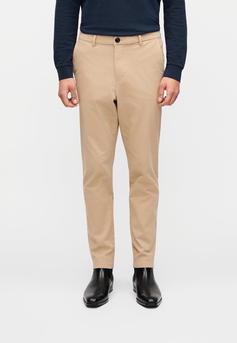 Μπεζ chino παντελόνια με ίσια γραμμή, διακοσμημένα με κουμπιά και θηλιές ζώνης, σε συνδυασμό με μαύρα μποτάκια αστραγάλου. Λείο βαμβακερό ύφασμα, λιτός σχεδιασμός.