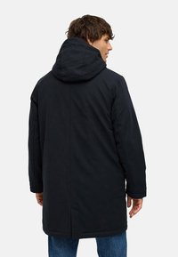 Donker gewatteerde parka met een gladde textuur, met een rechte silhouet, rugventilatie en lange mouwen. Gecombineerd met blauwe jeans.