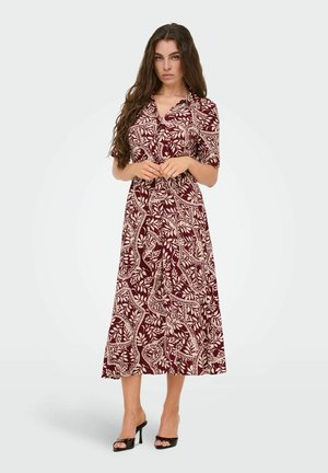 Žena s dlhými vlasmi oblečená v bordovo-béžových midi šatách s kvetinovým vzorom a čiernych sandáloch na podpätku s otvorenou špičkou, stojaca so zloženými rukami.