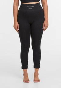 Zwarte leggings met een hoge tailleband, die een subtiele textuur en logo op de taille heeft. Materiaal lijkt rekbaar en figuurvormend.