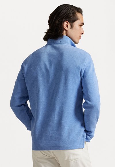 Pullover azul claro com um colarinho alto, tecido texturizado e fendas laterais na bainha. Tem mangas longas e um corte descontraído.