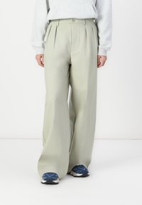 Pantalons taille haute vert clair avec des plis, en tissu lisse. Associés à des baskets bleu foncé. Le modèle se tient debout, les mains sur les côtés.