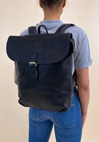 Una persona che indossa jeans blu e una maglietta grigia porta sulla schiena uno zaino in pelle nera con chiusura a fibbia.