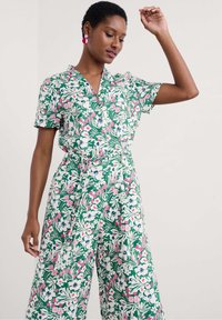 Zaļš ziedu kombinezons ar īsām piedurknēm, jostu viduklī un dzīvīgu rozā un zilā ziedu rakstu uz krēma krāsas fona.