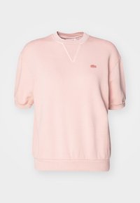 Jasnoróżowa bluza wykonana z miękkiego materiału, z prążkowanym dekoltem i krótkimi rękawami. Na piersi znajduje się małe haftowane logo.