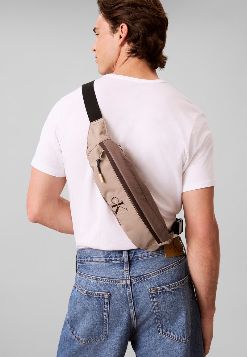 Homme portant un sac bandoulière beige et marron sur un t-shirt blanc et un jean bleu, vu de dos sur fond gris.