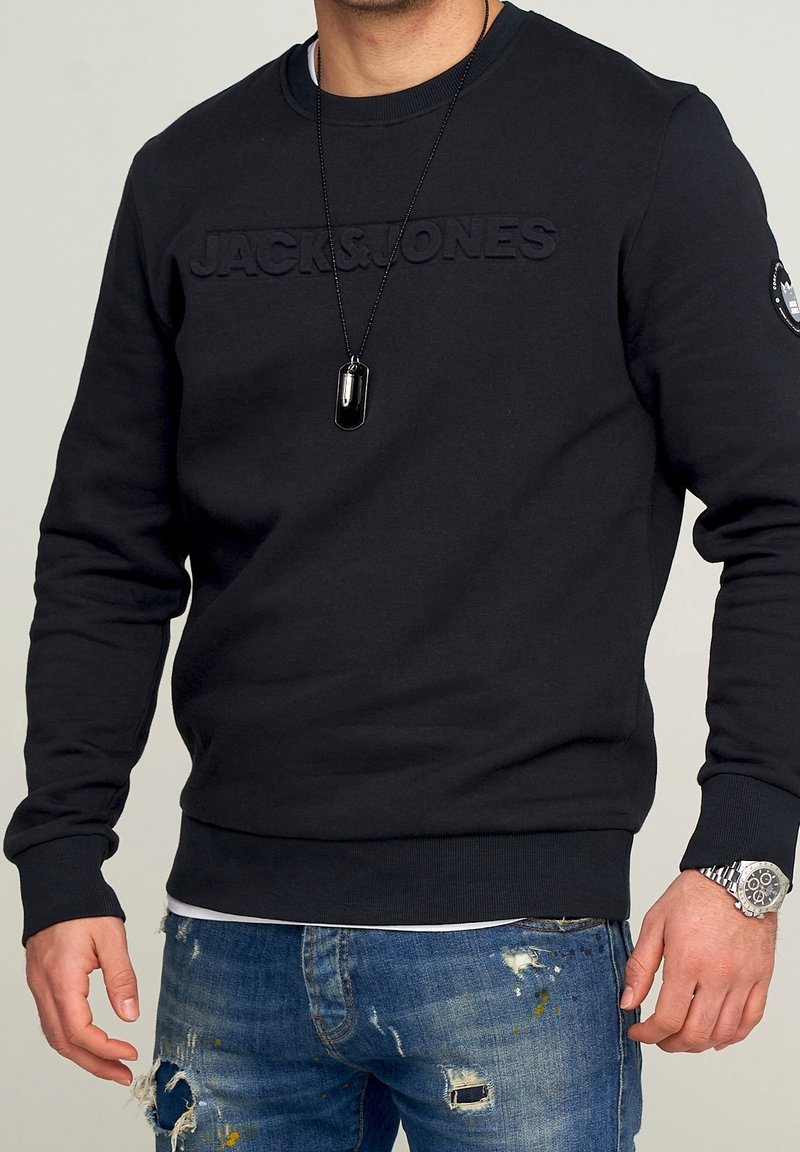Felpa nera con collo a giro e logo in rilievo "JACK & JONES", abbinata a jeans blu usurati e un orologio argentato.