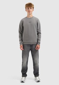 Tienerjongen staat, gekleed in een grijze sweatshirt met de tekst "BALLIN'", vervaagde zwarte jeans en witte sneakers tegen een effen lichte achtergrond.