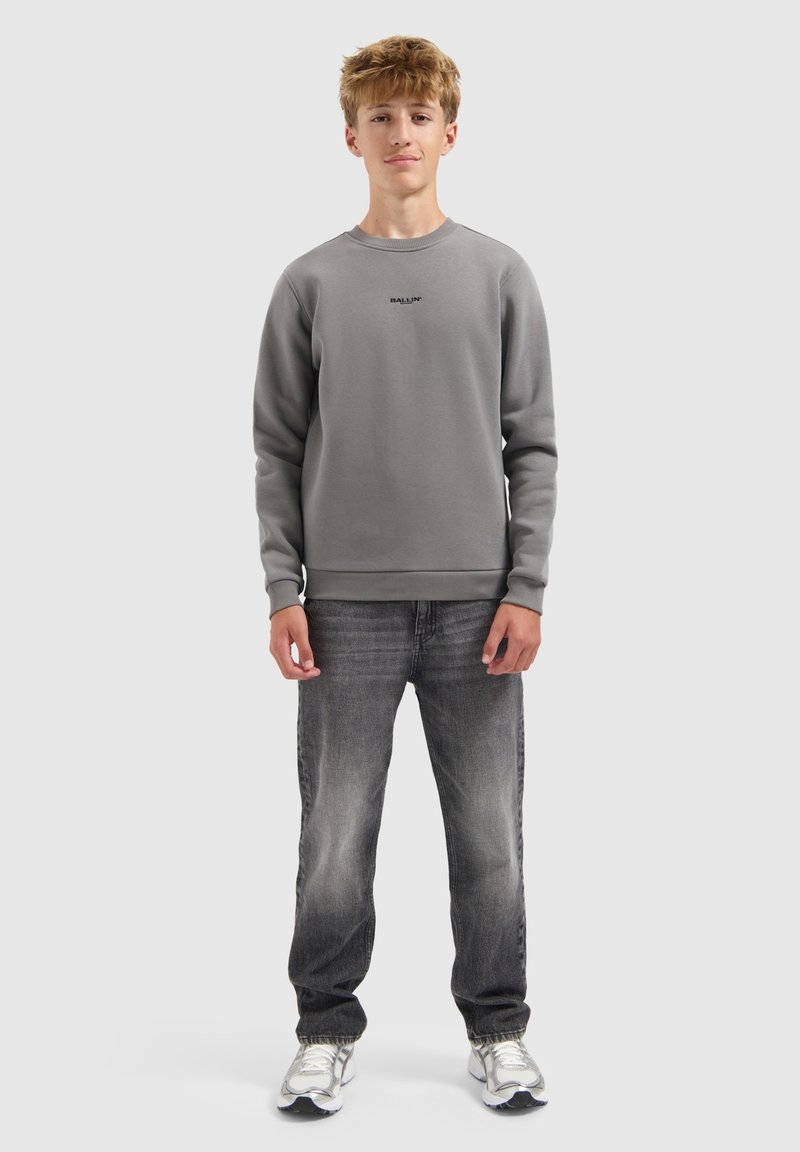 Tienerjongen staat, gekleed in een grijze sweatshirt met de tekst "BALLIN'", vervaagde zwarte jeans en witte sneakers tegen een effen lichte achtergrond.