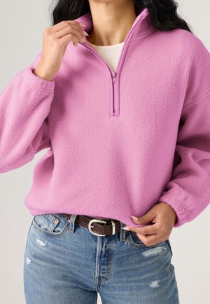 Femme portant un pull polaire rose avec une fermeture éclair sur la moitié, un haut beige clair en dessous, un jean bleu déchiré et une ceinture marron avec une boucle ovale.