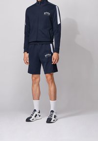 Chándal deportivo de la Marina con chaqueta de cremallera y pantalones cortos. Presenta rayas blancas en los lados y un logo. Se lleva con calcetines blancos y zapatillas deportivas.