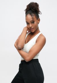 Baltas tankinis marškinėlis su V formos iškirpte, derinamas su juodomis sportinėmis kelnėmis. Modelis pozuoja sukryžiavęs rankas, demonstruodamas žaismingą šukuoseną.