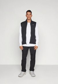 Črna flis brezrokavnik čez belo srajco z dolgimi rokavi, kombinirana s črnimi hlačami in sivimi športnimi copati. Preprost dizajn, brez vidnih vzorcev.