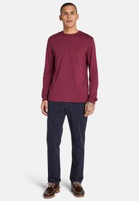 Langärmliges bordeauxrotes Hemd mit rundem Ausschnitt und kleinem Logo auf der Brust, kombiniert mit navyblauen Hosen und braunen Schuhen.