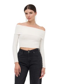 Top corto bianco a spalle scoperte in tessuto a costine con maniche lunghe, indossato con jeans neri a vita alta, caratterizzato da una texture liscia e un design aderente.