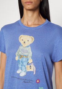 Modré tričko s grafikou medvídka v zeleném svetru a džínách, držícího košík. Měkký materiál s kulatým výstřihem a krátkými rukávy.