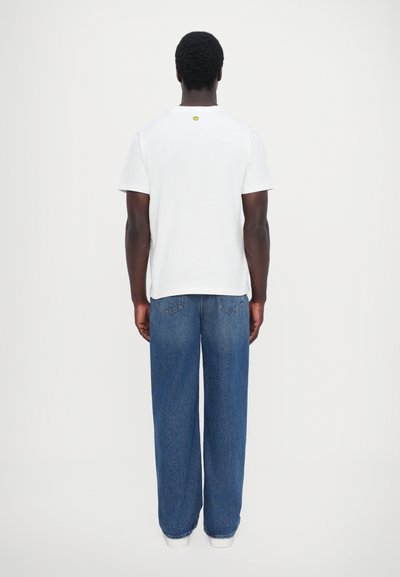 T-shirt en coton blanc à manches courtes, avec un petit accent jaune au dos. Porté avec un jean bleu ample.