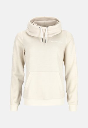 En kvinde står iført en beige hættetrøje og matchende sweatpants med hvide sneakers foran en ensfarvet baggrund.