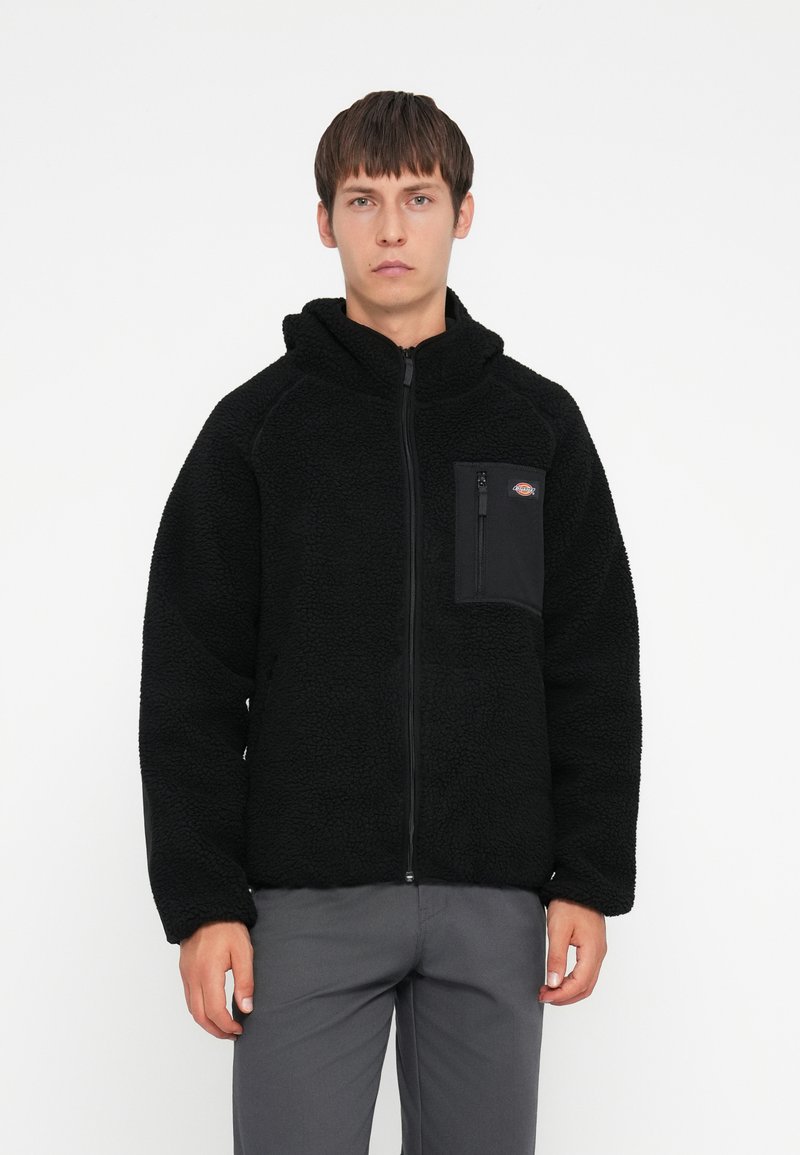 Veste en polaire noire avec une capuche, dotée d'une fermeture éclair et d'une poche sur la poitrine gauche en tissu noir. La veste a une finition douce et texturée.