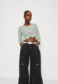 Hellgrünes gehäkeltes Crop-Top mit gewelltem Saum und offenem Strickdesign, kombiniert mit lockeren schwarzen Cargo-Hosen mit Reißverschlüssen und Taschen.
