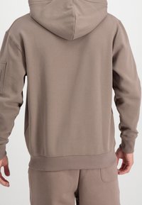Sweat à capuche en tissu taupe. Dispose d'un ourlet et de poignets côtelés, avec une poche latérale et un détail de couture minimaliste sur l'épaule.