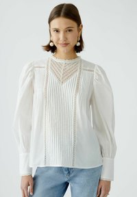 White blouse with long puff sleeves, pleated front, lace accents, and scalloped neckline, paired with light blue jeans.