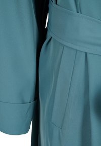 Primo piano di un vestito di stoffa teal con tasca, dettaglio della cintura e polsino della manica piegato.