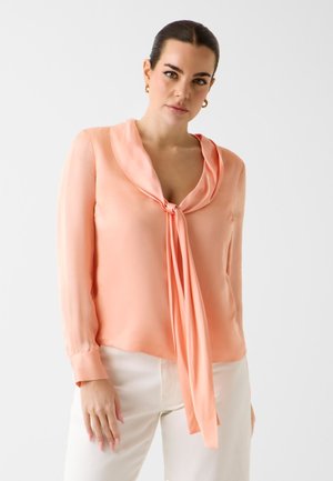 Femme aux cheveux foncés en chignon bas, portant des boucles d'oreilles anneaux dorés, un chemisier pêche avec un lien à l'avant, et un pantalon blanc sur un fond clair uni.