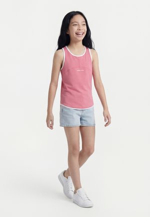 Dívka se usmívá a kráčí, oblečená v růžovém tílku, světlých džínových šortkách a bílých teniskách na bílém pozadí.