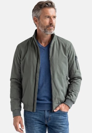 Mann mit grauem Haar und Bart, der eine grüne Jacke mit Reißverschluss über einem blauen Pullover und Jeans trägt, schaut seitlich mit einer Hand in der Tasche.