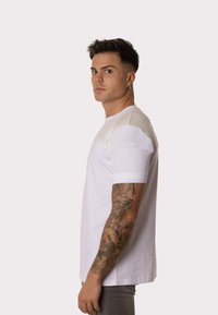Camiseta blanca de manga corta con acentos texturizados en los hombros, escote redondo y corte relajado; el modelo posa de lado mostrando el detalle de las mangas.