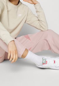 Beżowa bluza Nike, różowe dopasowane spodnie dresowe z bocznymi akcentami, białe skarpetki oraz białe sneakersy Nike Air z zielonymi i czerwonymi detalami.