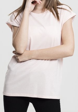 Jeune femme portant un t-shirt rose clair à manches courtes et un pantalon noir, bras croisés et main touchant son menton, sur un fond uni.