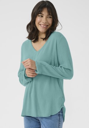 Blouse teal à manches longues avec un décolleté en V, texture douce et coupe décontractée. L'ourlet est légèrement courbé avec des fentes latérales pour faciliter les mouvements.