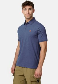 Man die een blauw poloshirt met korte mouwen en kaki cargobroek draagt, staat tegen een simpele witte achtergrond, kijkt recht vooruit.