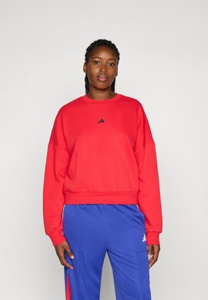 Red sweatshirt with a crew neck and long sleeves, featuring a small black Adidas logo on the chest. Worn with blue pants.