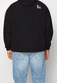 Svart huva jacka med en logotyp på ryggen och ett vitt rutmönster längs ärmarna. Mjuk material, avslappnad passform och långa ärmar.