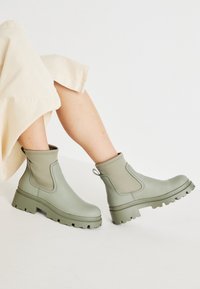Botas de tobillo verdes con un acabado suave, paneles laterales elásticos y una suela gruesa. Presenta una lengüeta en la parte trasera y una textura estriada en la suela.