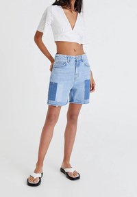 Shorts en denim bleu clair avec un design patchwork comprenant différentes nuances de bleu. Ourlet effiloché, poches standard et taille haute. Associés à un crop top blanc.