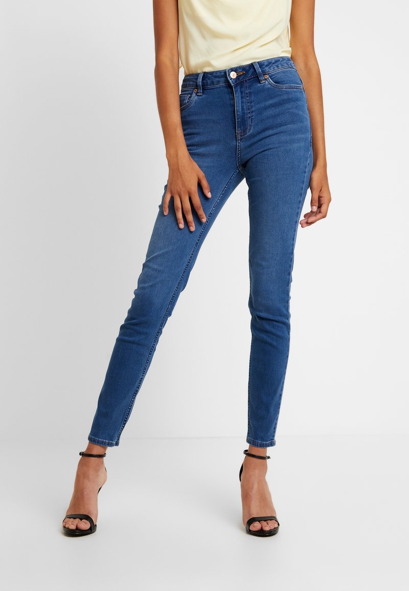 Jeans de mezclilla azul ajustados con una cintura de media altura, diseño de cinco bolsillos y un ligero desvanecimiento. Combinados con tacones negros de tiras para crear contraste.