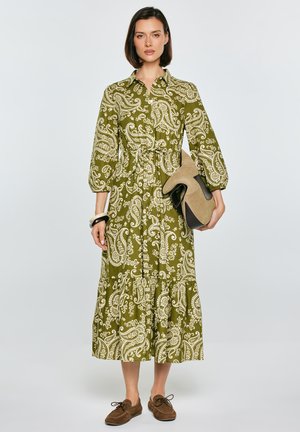 Kobieta w oliwkowo-zielonej sukience midi z białym wzorem paisley, trzymająca beżowo-czarne botki, stojąca o neutralnym wyrazie twarzy.