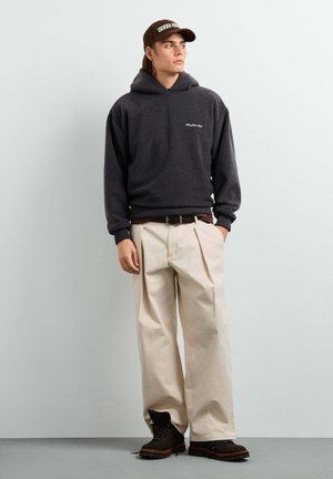 Junger Mann trägt schwarzen Fleece-Hoodie, beige weite Hosen, schwarze Stiefel und eine braune Baseballkappe und schaut seitlich vor einem einfarbigen Hintergrund.