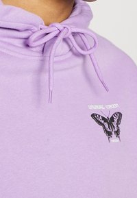 Felpa con cappuccio viola realizzata in tessuto morbido, dotata di chiusura con coulisse e una grafica di una farfalla nera con il testo "Visione Insolita".